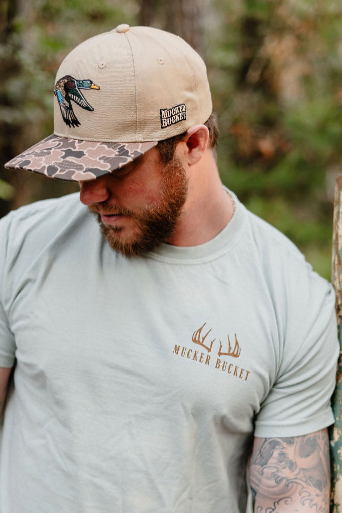 A bearded man wearing a beige Mucker Bucket hat with a bird patch and a Buck Season Premium Tee by Mucker Bucket, featuring an antler logo, stands outdoors looking down with trees in the background.