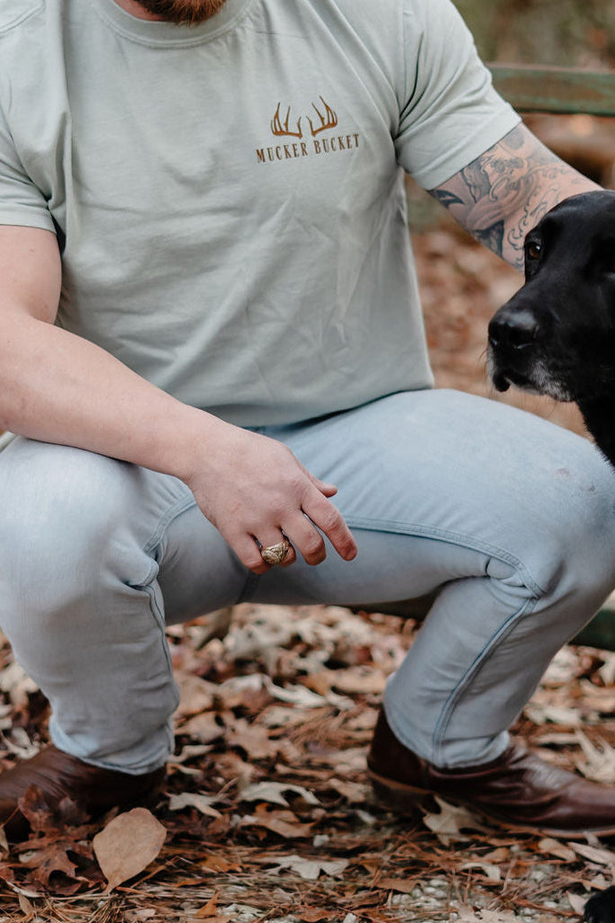Wearing a light gray Mucker Bucket Buck Season Premium Tee and light jeans, a tattooed person squats outdoors on dry leaves, extending an arm to a black dog. Only part of their face and the dog’s head are visible.
