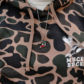 A close-up shows someone in a USA-made camouflage hoodie with a flying duck design, featuring shades of green, brown, and black. Over the hoodie, they wear Mucker Buckets Wood Duck Necklace—a silver stainless steel chain with a wood duck pendant.