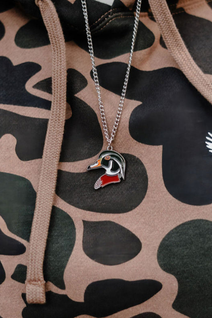 A close-up shows someone in a USA-made camouflage hoodie with a flying duck design, featuring shades of green, brown, and black. Over the hoodie, they wear Mucker Buckets Wood Duck Necklace—a silver stainless steel chain with a wood duck pendant.