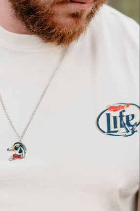 A close-up shows a bearded man wearing the Mucker Bucket Mallard Life Premium Tee, garment dyed in a relaxed fit with a Miller Lite logo, and sporting a silver duck-shaped pendant. Only the lower half of his face is visible.