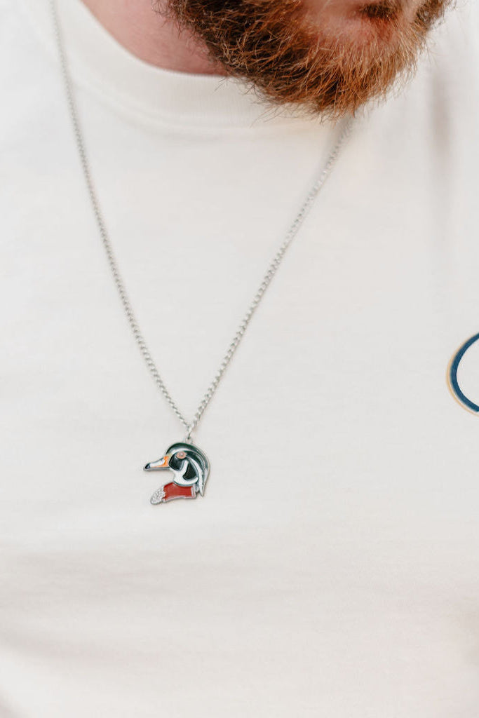 Close-up of a person with a reddish beard wearing a white shirt featuring a Life logo and the Mucker Bucket Wood Duck Necklace with stainless steel chain.