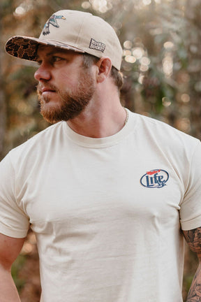 A bearded man in a beige Mucker Bucket Mallard Life Premium Tee with a Lite logo and a tan cap stands outdoors, glancing sideways as tall trees and soft sunlight fill the blurred background.