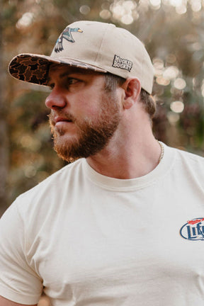 A bearded man wearing the Mucker Bucket Mallard Old School Duck Bucket hat and a white T-shirt with a small logo stands outdoors in soft, natural light during hunting season.