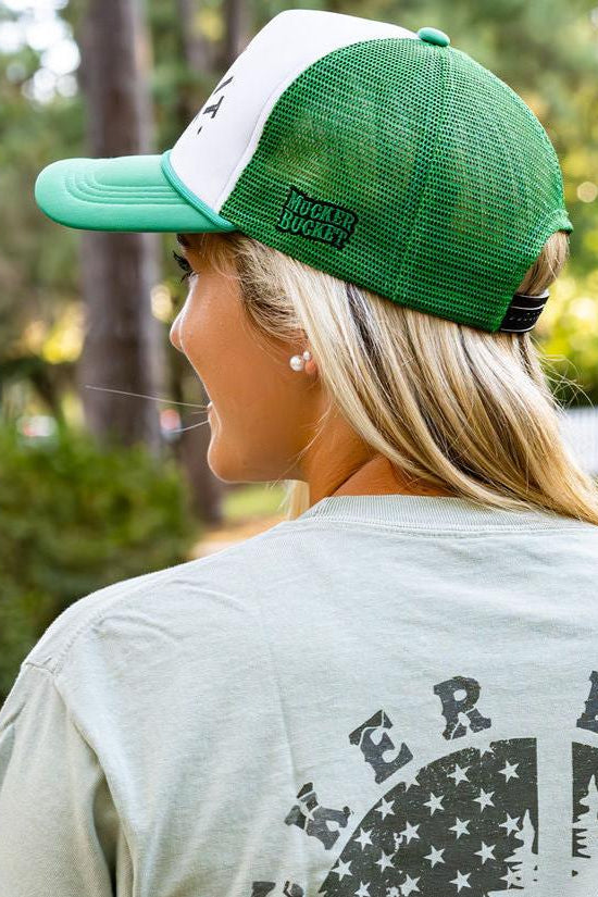 A person with long blonde hair is seen from behind, wearing a green and white Duck It Green Trucker Hat by Mucker Bucket and a light green shirt featuring a high-quality screen-printed circular logo on the back. Trees and a white fence blur in the background.