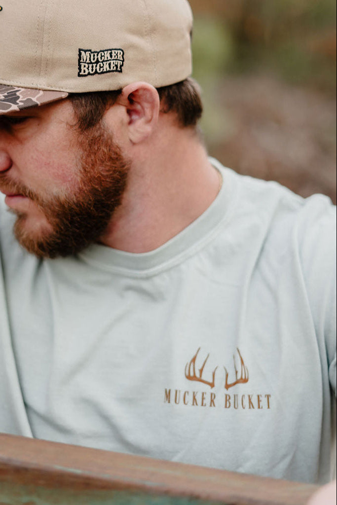 A bearded man wears a beige cap and a light gray Buck Season Premium Tee by Mucker Bucket, featuring “Mucker Bucket” text with deer antler graphics. The blurred background highlights this stylish hunting shirt.