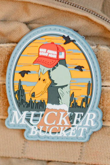 The Smart Duck PVC Patch by Mucker Bucket is a 4x4 3D PVC patch showing a duck in a red "Make Great Again" hat, backed by a desert scene with birds. Velcro tear-away backing allows for easy attachment. “MUCKER BUCKET” appears at the bottom.