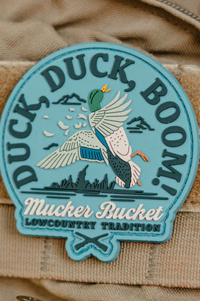 The Duck Duck Boom PVC Patch by Mucker Bucket is a round blue patch featuring a flying mallard, “Duck Duck Boom!” at the top, and “Mucker Bucket, Lowcountry Tradition” at the bottom with clouds and grass in the background.