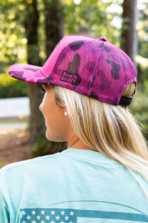 A person with long blonde hair wears a Mucker Bucket "Duck It" Pink Camo Trucker Bucket hat with a mesh back and patch, paired with a light blue shirt, standing outside among trees in the sunlight.