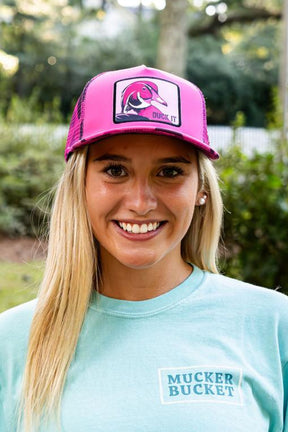 A smiling young woman wears a "Duck It" Pink Camo Trucker Bucket by Mucker Bucket and a light blue T-shirt, standing outdoors with greenery and trees softly blurred in the background.