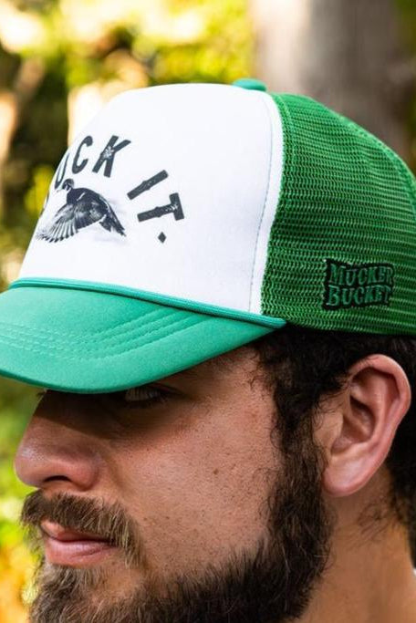 A man wearing the Duck It Green Trucker Hat, by Mucker Bucket, with a mesh back and BUCK IT text alongside a flying bird graphic on the front. The side features a green MUCKER BUCKET logo. This standout hat contrasts against blurred green foliage.