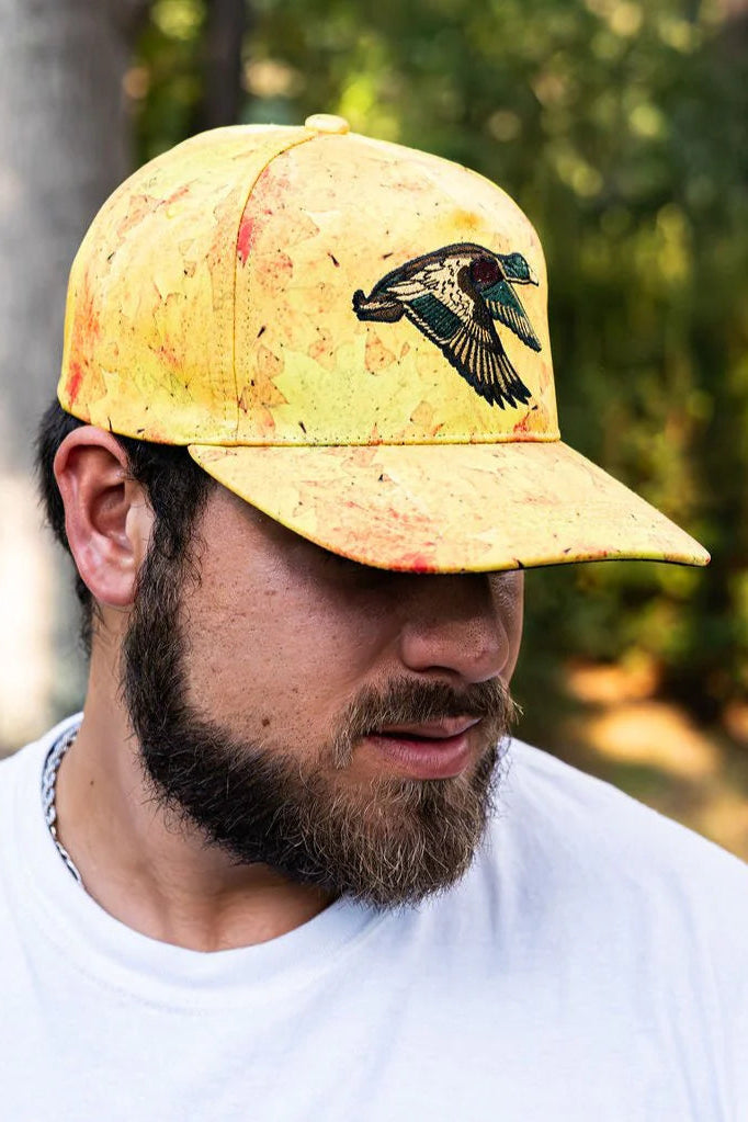 A bearded man in a white t-shirt and yellow Mucker Bucket Golden Leaf Camo Duck Bucket hat with a bird design looks down, set against a blurred outdoor scene of trees and greenery, suggesting serene duck hunting adventures.