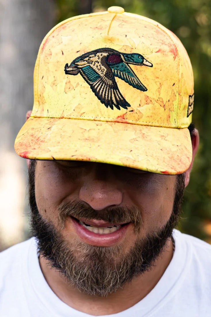 A bearded man stands outdoors, smiling beneath a Golden Leaf Camo Duck Bucket from Mucker Bucket. The blurred, lush green background enhances his relaxed demeanor.