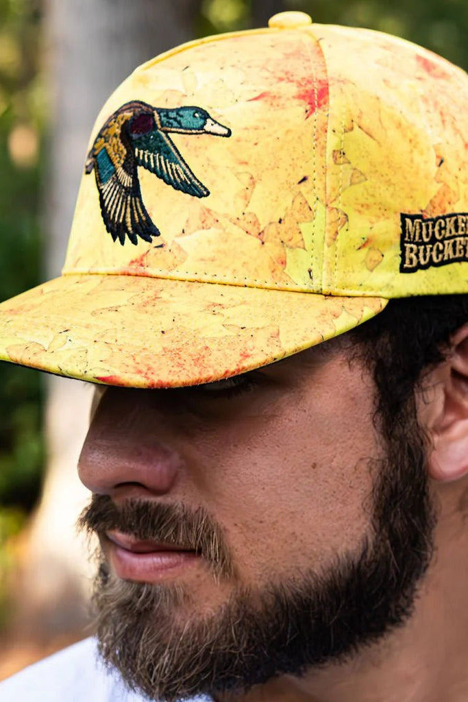 A bearded man wears the Golden Leaf Camo Duck Bucket hat by Mucker Bucket, with a bird design and logo, reminiscent of duck hunting adventures. Blurred green foliage forms his backdrop.