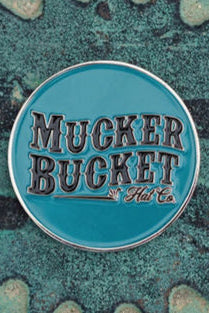 A round teal pin adorned with Mucker Bucket in bold black letters against a textured background with irregular shapes and a subtle duck design, named Duck, Duck, Boom Lucky Huntin Coin.