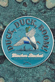 Round pin badge featuring a flying duck and the text Duck, Duck, Boom! with Winchester Buckshot below. The teal background adds texture, reminiscent of Mucker Buckets Duck, Duck, Boom Lucky Huntin Coin.