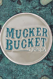 A round metal emblem with raised blue-green Mucker Bucket Hat Co. lettering on a silver background resembles the Chasin Tail Lucky Huntin Coin from Mucker Bucket. It rests on a textured, weathered green surface.