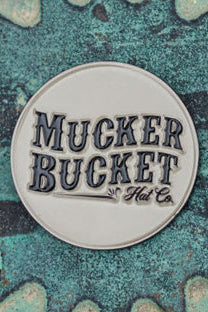 A gray patch with bold black Mucker Bucket Hat Co. text rests on a textured teal backdrop with gear patterns, resembling the Got One Lucky Huntin Coin. Why not get one for yourself?.