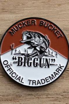 A circular emblem with a fish leaping from water resembles the Biggun Lucky Fishin Coin. The top half displays the Mucker Bucket brand in brown, while the bottom half is white, featuring the bold Biggun Official Trademark. The background has a wooden texture.