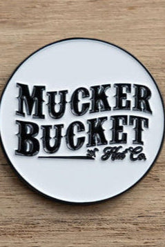 A round pin with Mucker Bucket in bold black text on a white background rests on wood, like the Biggun Lucky Fishin Coin washed ashore, awaiting adventures.