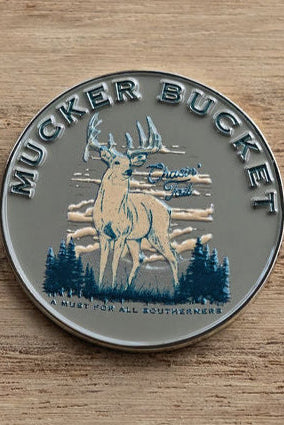 The Mucker Bucket Chasin Tail Lucky Huntin Coin features a round metal pin with a detailed buck, antlers, trees, and clouds. The text A Must for All Southerners rests on a wooden backdrop, embodying the Southern spirit.
