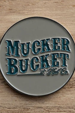 A round metallic badge embossed with Mucker Bucket in stylized blue-green lettering against a rustic wooden backdrop, like the Chasin Tail Lucky Huntin Coin, ready to bring fortune on your next hunt.