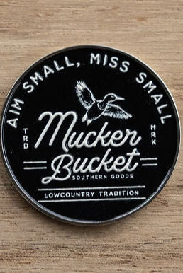 The Aim Small, Miss Small Lucky Huntin Coin by Mucker Bucket features a round black design with white text and a flying duck illustration. Below, it reads, A Lowcountry Tradition. This unique piece is an ideal addition to any hunting enthusiasts collection.