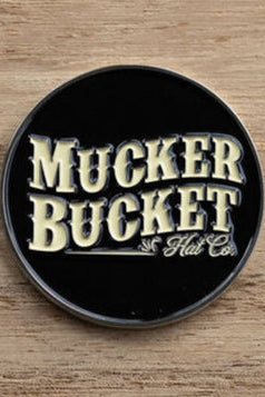 A round, black pin with Mucker Bucket in bold, yellow letters rests on a wooden surface, much like the Aim Small, Miss Small Lucky Huntin Coin waiting to be found.