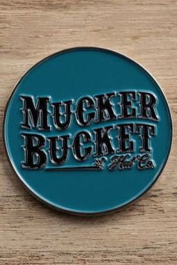 A circular teal metal pin embossed with Mucker Bucket Hat Co. lies on a wooden surface next to the Duck, Duck, Boom Lucky Huntin Coin charm.