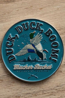 A round blue pin badge features a flying duck with DUCK, DUCK, BOOM! above and Mucker Bucket below, reminiscent of the charm of the Mucker Bucket brands Duck, Duck, Boom Lucky Huntin Coin.