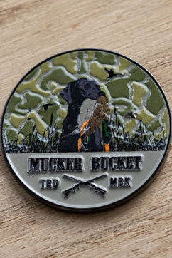 The Got One Lucky Huntin Coin by Mucker Bucket features a black dog with a bird in its mouth on a camo background. It says MUCKER BUCKET with crossed rifles below and TRD MRK on the sides, all resting on a wooden surface, capturing the essence of luck in hunting.