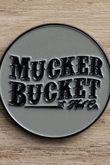This Got One Lucky Huntin Coin by Mucker Bucket is a round metal badge featuring embossed letters against a wooden backdrop with a glossy finish, exuding charm and serendipity.