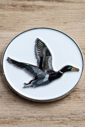 The OG Duck Lucky Huntin Coin by Mucker Bucket features a detailed illustration of a flying duck with a green head and gray body on a white background. The round pin elegantly rests on wood, ready to bring you luck in your next adventure.