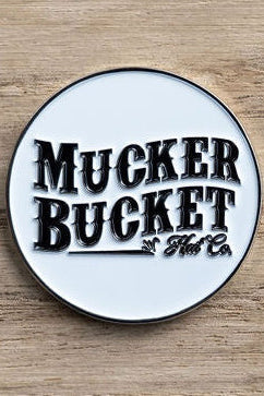 A circular logo with a white background displays Mucker Bucket in bold black letters, reminiscent of an OG design. The logo elegantly rests on a wooden surface, matching the style of the OG Duck Lucky Huntin Coin.
