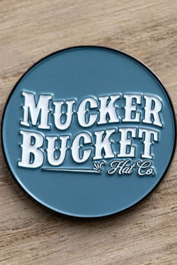 A blue, round pin sits on a wooden surface, featuring white text with Mucker Bucket branding in a vintage style, echoing the Smart Duck Lucky Huntin Coin era.