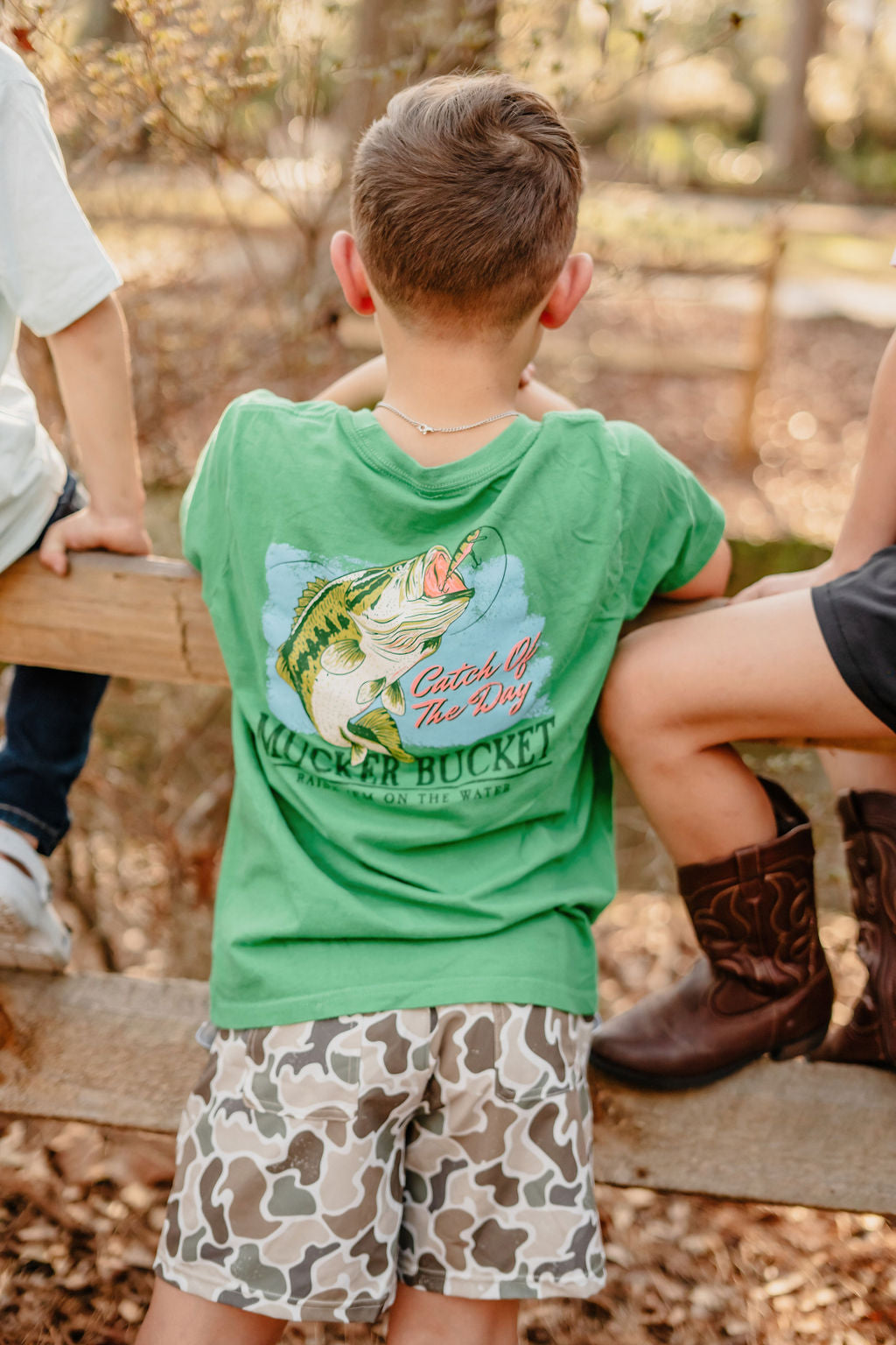 A child with short hair, wearing a green Catch of the Day fish graphic tee and Mucker Buckets Youth Performance Athletic Shorts in camo, is sitting on a fence. Another person in boots sits beside them outdoors among trees and a fence.