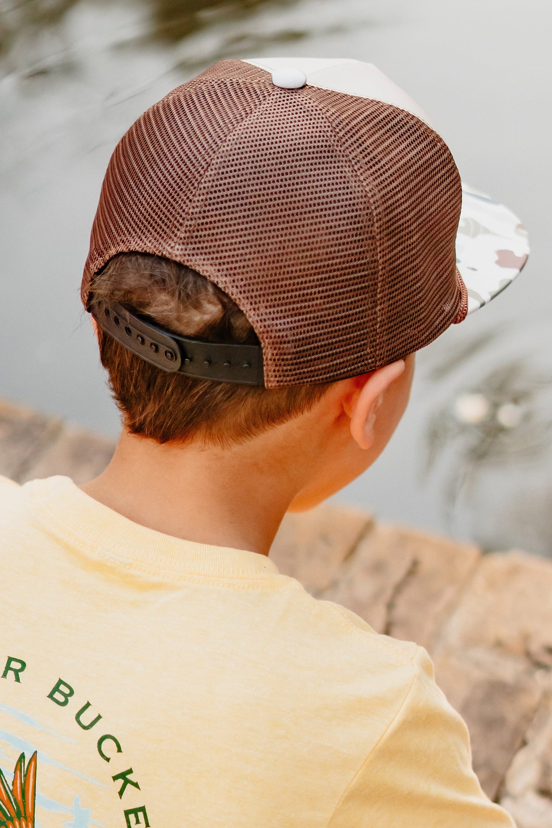 Person wearing a brown and white cap and yellow shirt with 'Mucker Buckets' logo by a body of water.
