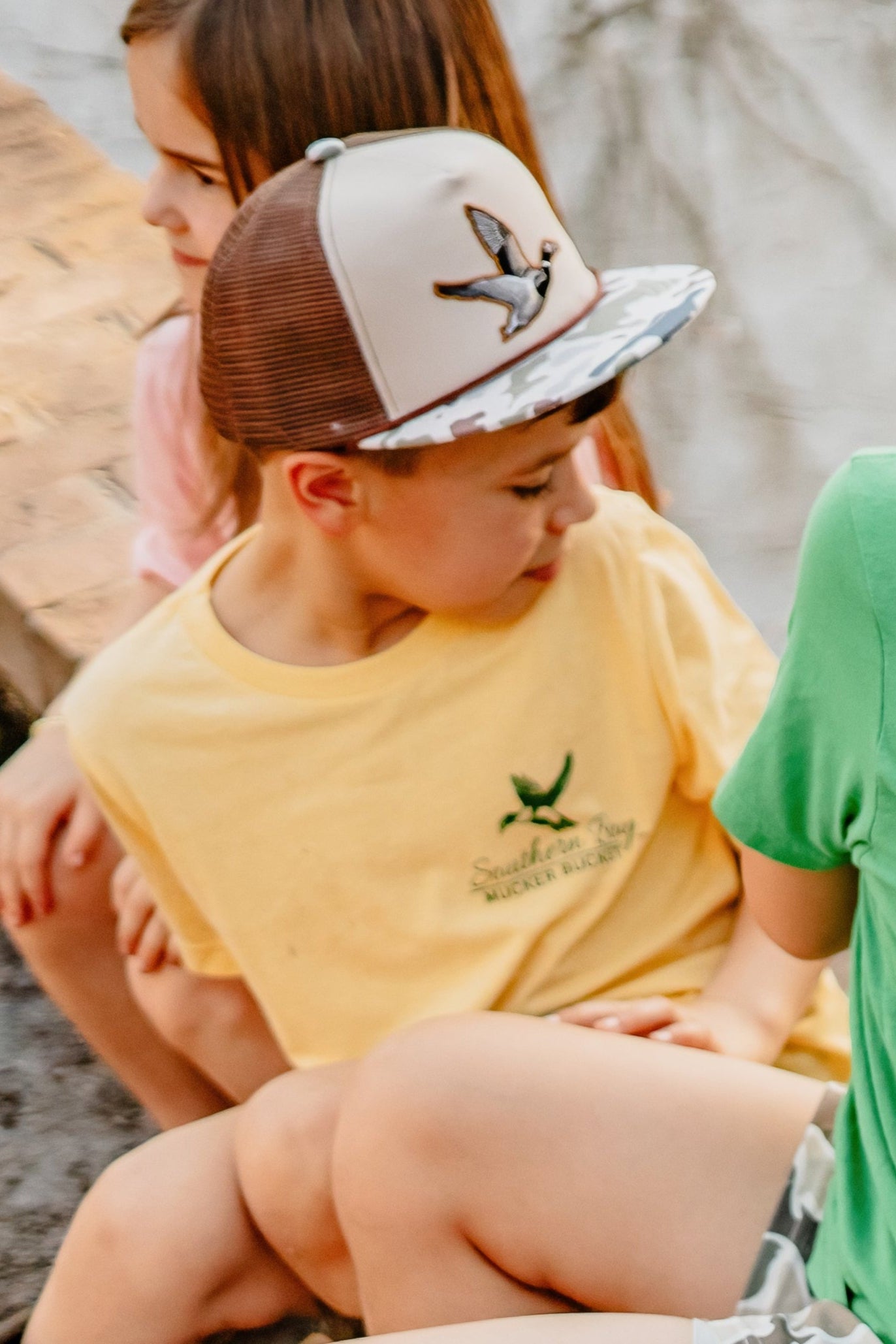 Children sitting outdoors, wearing yellow and green shirts with a logo.