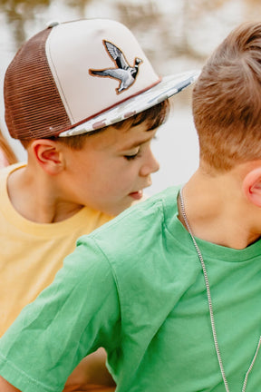 Two young boys stand close outdoors. One wears a yellow shirt and the Mucker Bucket Youth Camo Duck Bucket Hat, while the other sports a green shirt and silver chain. Their faces are partly visible.
