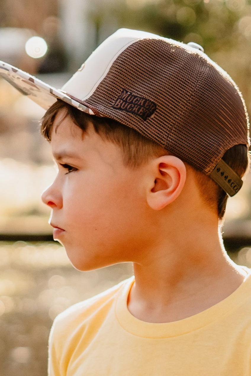 A young boy in a yellow shirt wears the Mucker Bucket Youth Camo Duck Bucket Hat, looking to the left in soft outdoor sunlight with a blurred background.