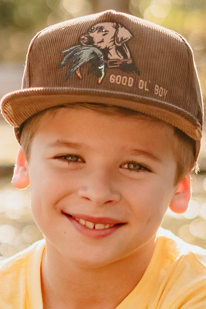 Young boy wearing a brown cap with a dog design and text, smiling outdoors.