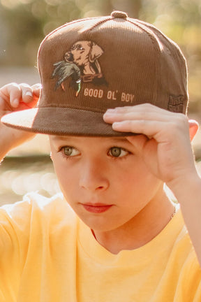 Child wearing a brown cap with a design and text, outdoors.