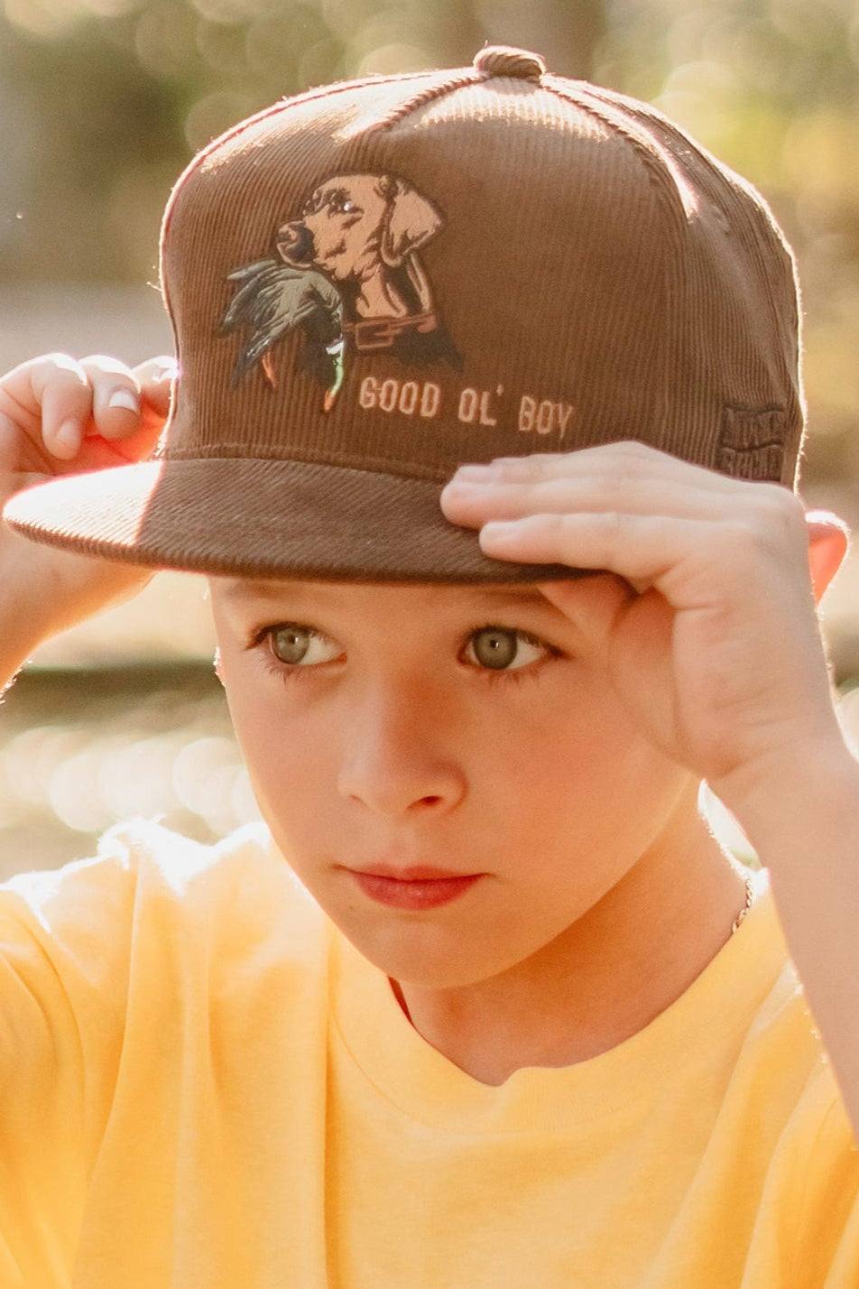 Child wearing a brown cap with a design and text, outdoors.
