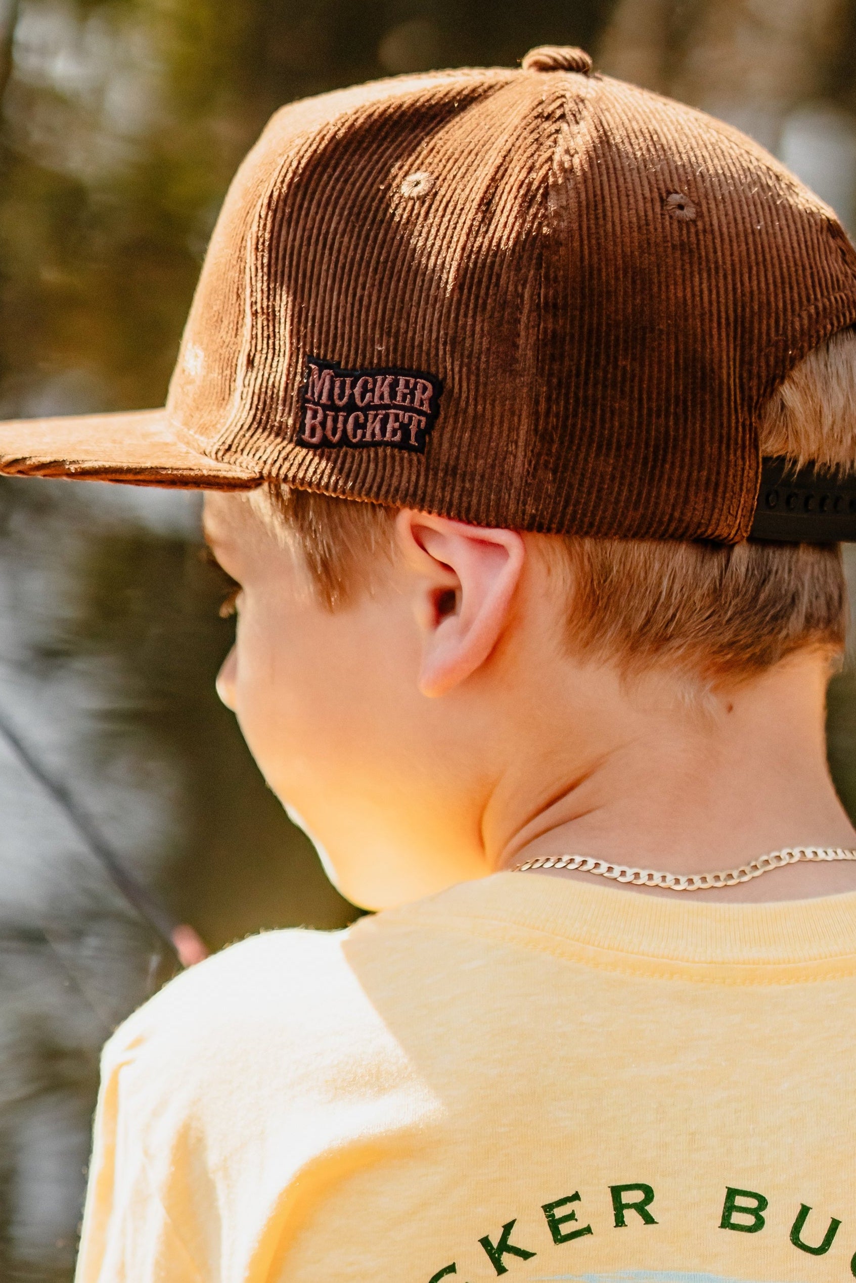A young boy stands outdoors with sunlight highlighting his hair, wearing the Mucker Bucket Youth Good Ol' Boy Corduroy Bucket Hat and a yellow T-shirt.