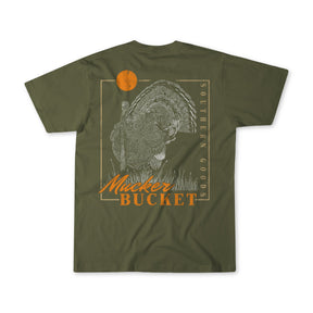 The Ghost Gobbler Premium Tee by Mucker Bucket is an olive green, garment-dyed shirt with a detailed turkey, orange sun, and Southern Goods, Mucker Bucket in white and orange on the back.