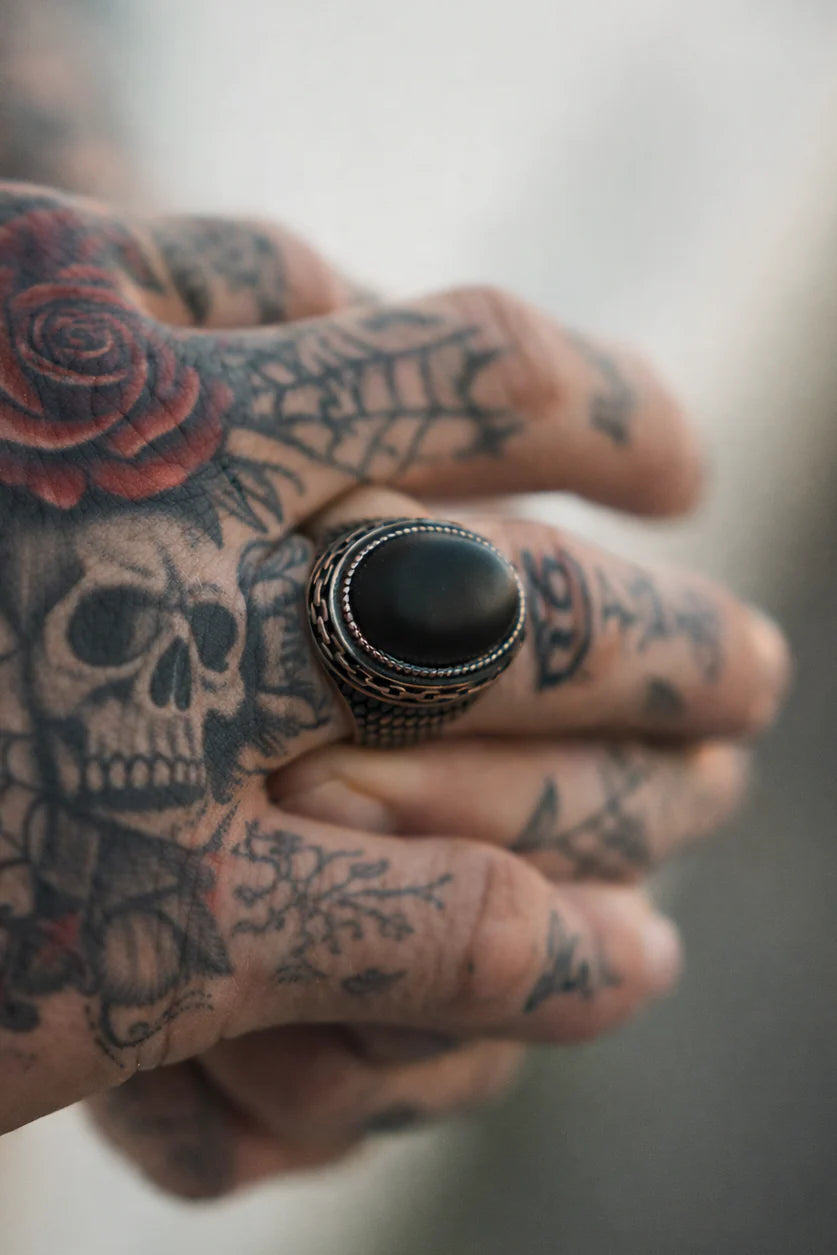 A tattooed hand adorned with a skull and rose design wears The Dark Current Ring by Relentless Betrayal—a large, handcrafted men's ring showcasing an oval black stone on the ring finger.