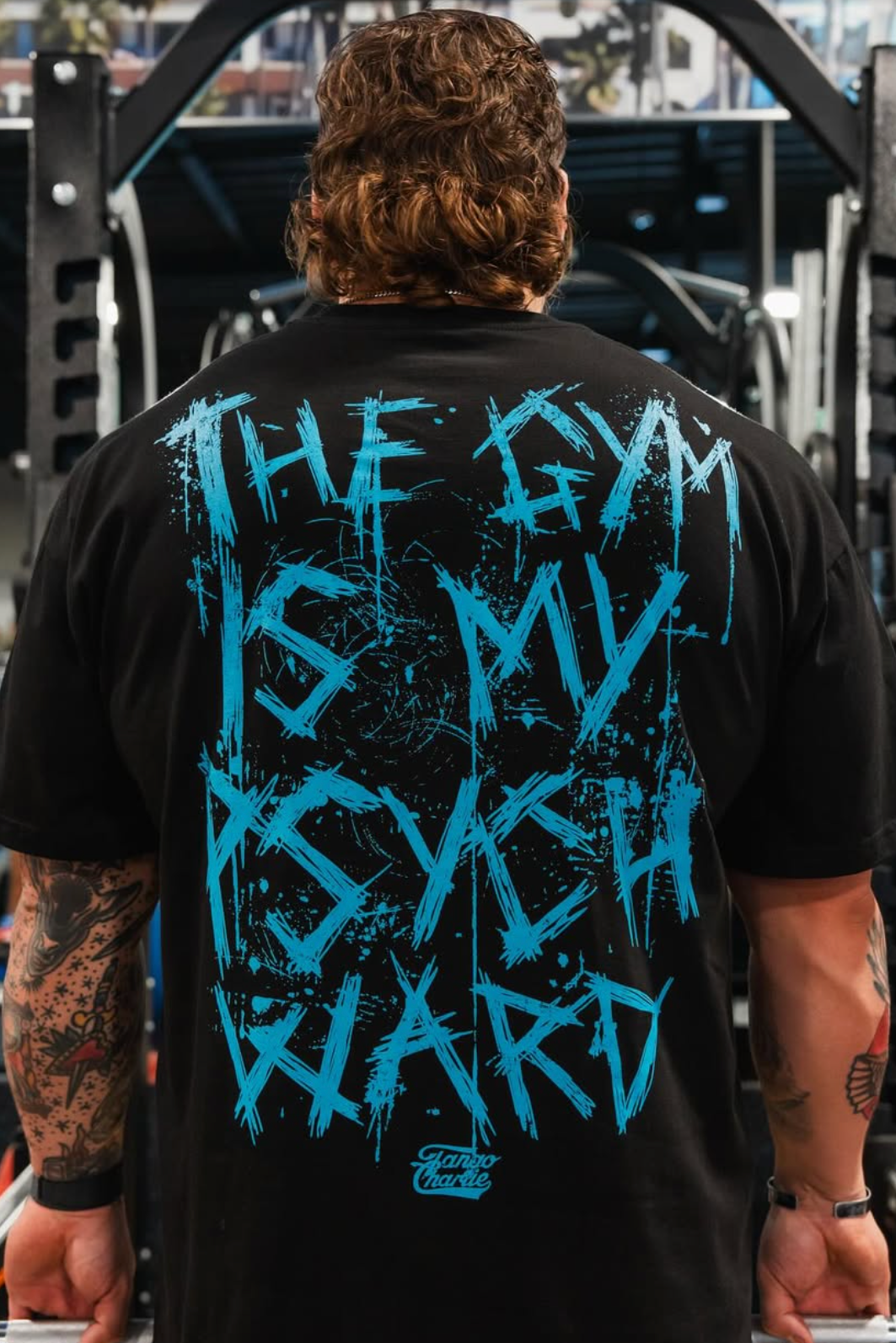 A person with tattooed arms is seen from behind in a gym, wearing the Tango Charlie Apparel Psych Ward - Premium Oversized Tee featuring blue graffiti text, THE GYM IS MY PSYCH WARD, with gym equipment and people in the background.
