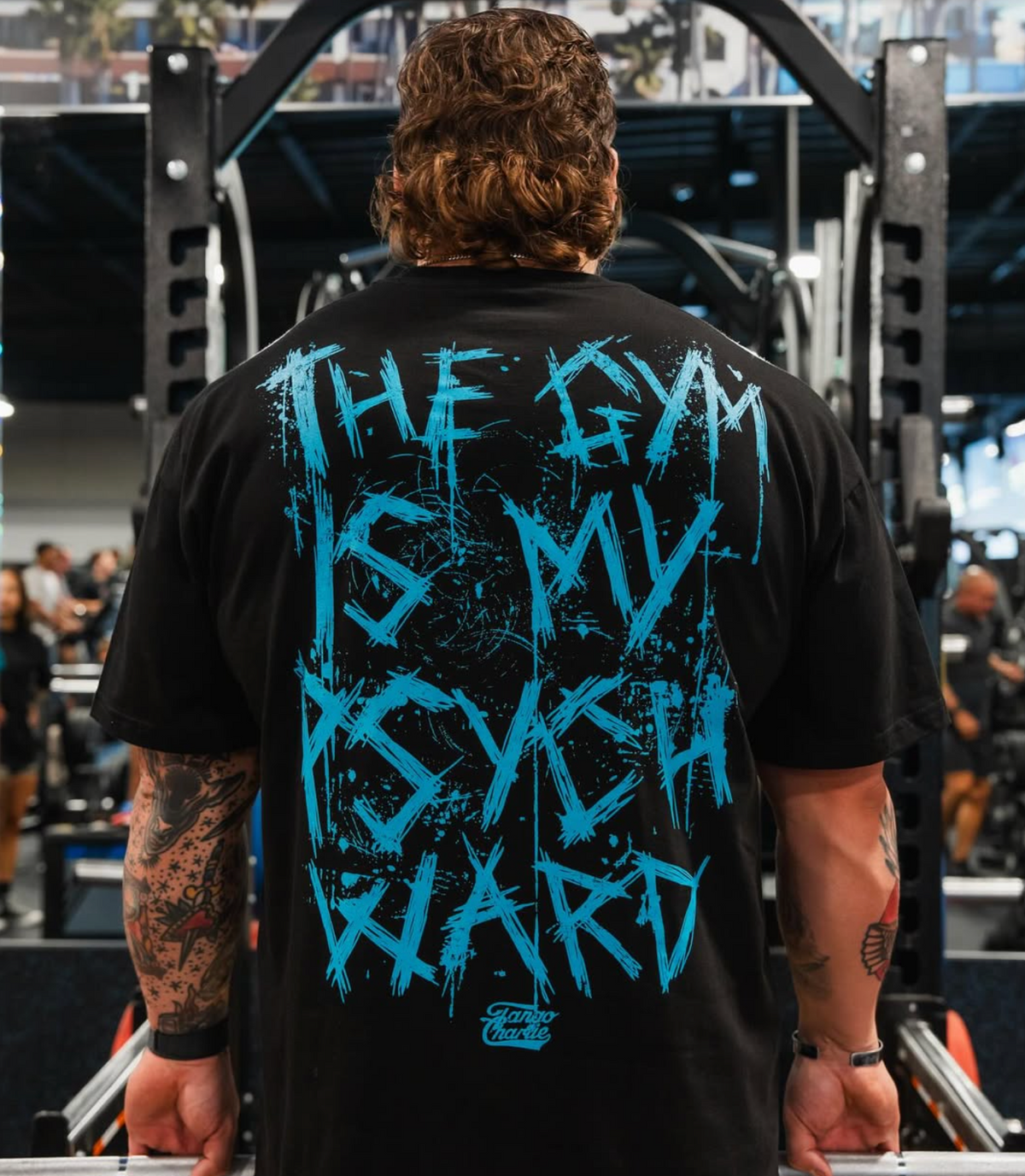 A person with tattooed arms is seen from behind in a gym, wearing the Tango Charlie Apparel Psych Ward - Premium Oversized Tee featuring blue graffiti text, THE GYM IS MY PSYCH WARD, with gym equipment and people in the background.