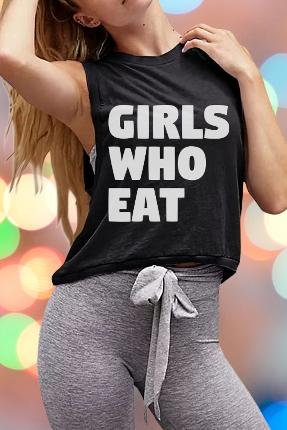 A woman models the Tango Charlie Apparel “Girls Who Eat” Crop Tank—a sleeveless black top emblazoned with bold lettering—paired with gray front-tie leggings, standing against a vibrant, blurred background for a confident and empowered look.