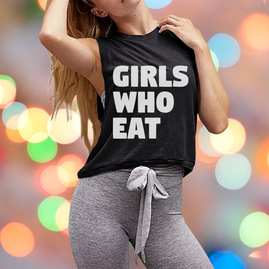 A woman wears the Girls Who Eat Crop Tank by Tango Charlie Apparel, featuring bold white text, paired with gray drawstring leggings. This comfy top pops against a backdrop of colorful, blurred lights.